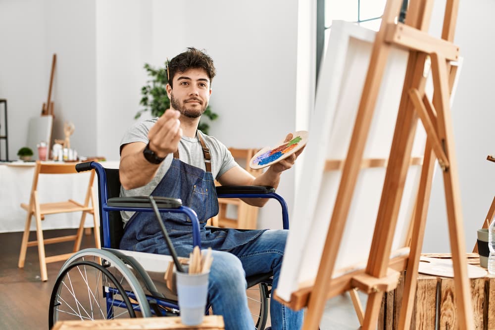 Young hispanic man sitting on wheelchair painting at art studio doing money gesture with hands